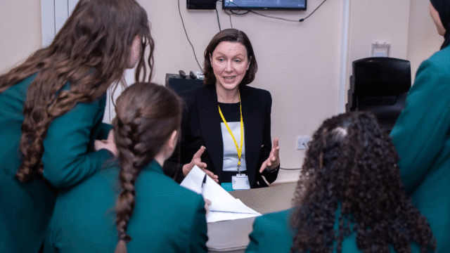 4 secondary school girls around a table facing away looking at a woman who us sat down on the other side of the table talking to them about her profession