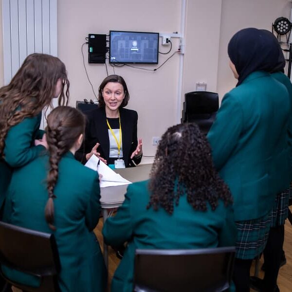 4 secondary school girls around a table facing away looking at a woman who us sat down on the other side of the table talking to them about her profession