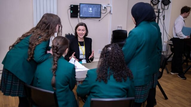 4 secondary school girls around a table facing away looking at a woman who us sat down on the other side of the table talking to them about her profession