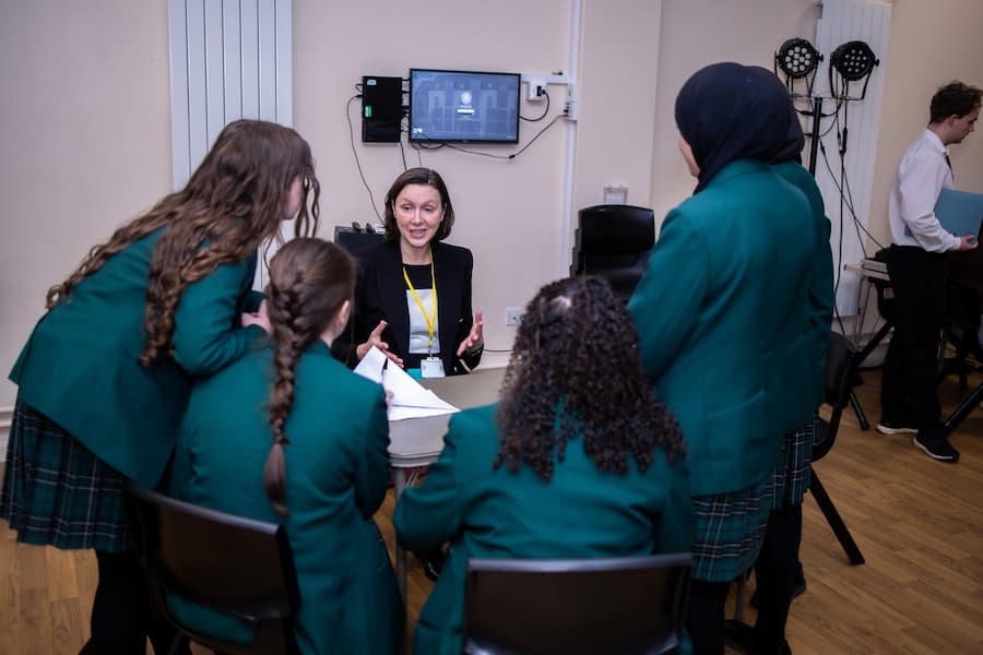 4 secondary school girls around a table facing away looking at a woman who us sat down on the other side of the table talking to them about her profession
