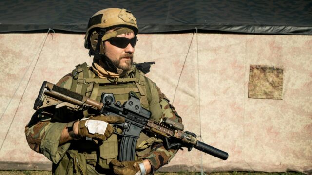 A soldier in full gear with an assault rifle in front of a tent