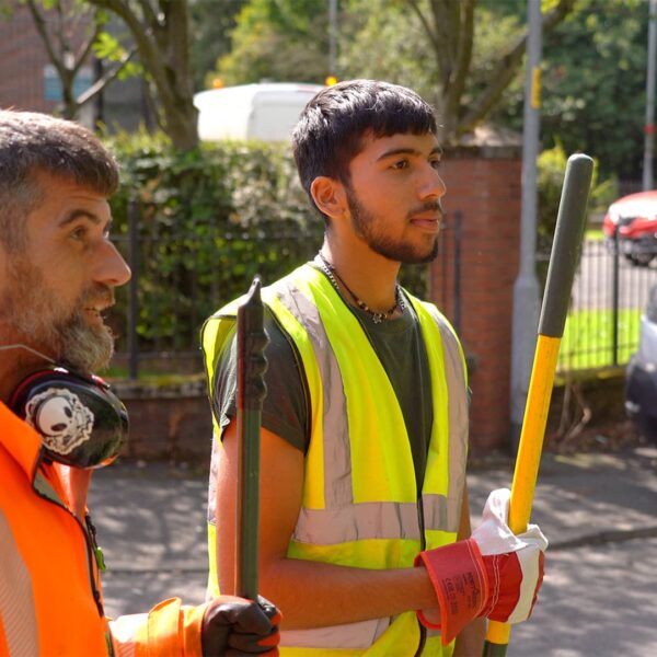 A young man on a work placement with Manchester City council's parks team. He is holding a gardening fork and speaking with his supervisor. They are both in high vis workwear.