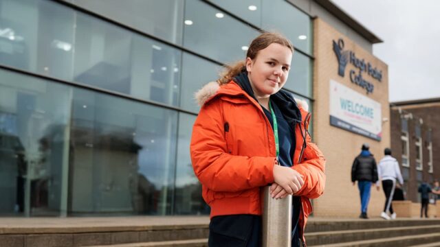 A young woman stood in front of Hugh Baird College in an orange coat, smiling