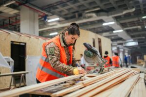 A woman in a hi-vis jacket in a workshop marking some wood before she cuts it with a buzzsaw
