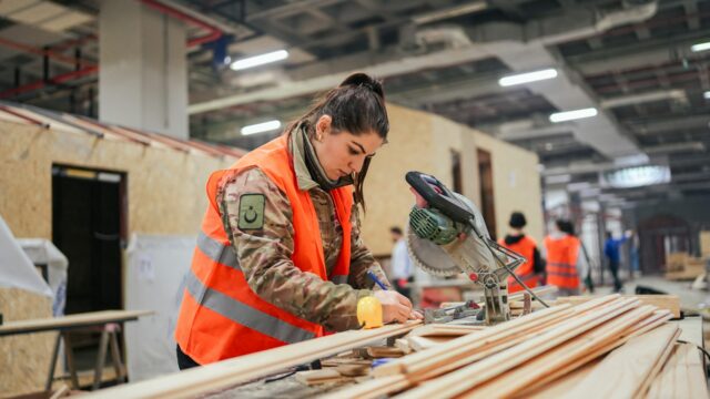 A woman in a hi-vis jacket in a workshop marking some wood before she cuts it with a buzzsaw