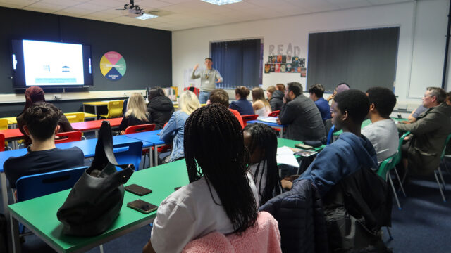 A classroom full of students looking towards a teacher who is demonstrating something at the front of the classroom