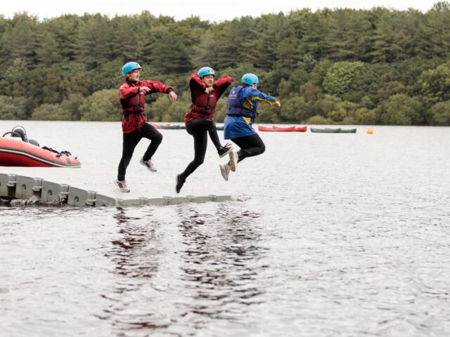 Three young men on an outward bound course are wearing life jackets and waterproofs. They are jumping into water with joy.