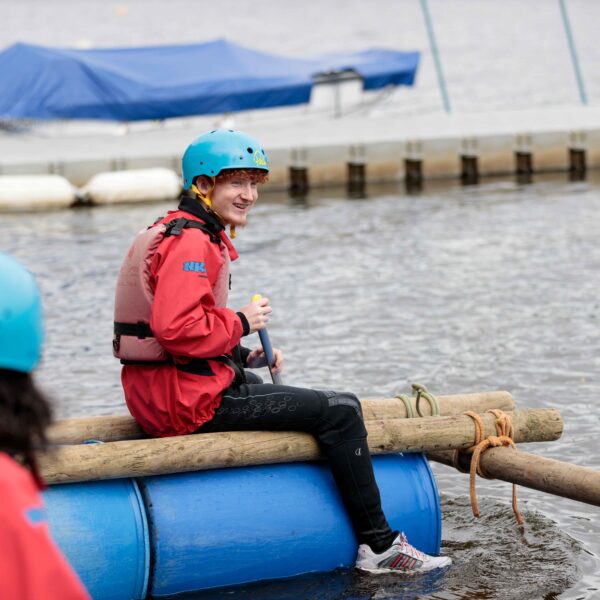 A young man is on a hand-made raft, made from barrels and wood. This is part of a construction activity course. he is on the raft on the water, with a helmet and lifejacket on, and he is smiling.