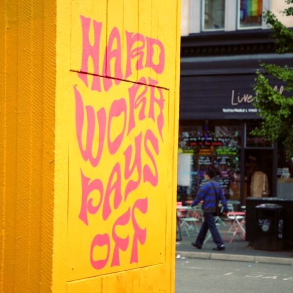 A piece of street art in Manchester. A plinth is painted in yellow, with pink psychadelic wording: "hard work pays off".