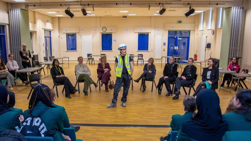 A group of professional women all in different uniforms sat in a horseshoe shape with one woman, who is wearing a hard hat and a hi-vis jacket stood in the middle talking to an audience