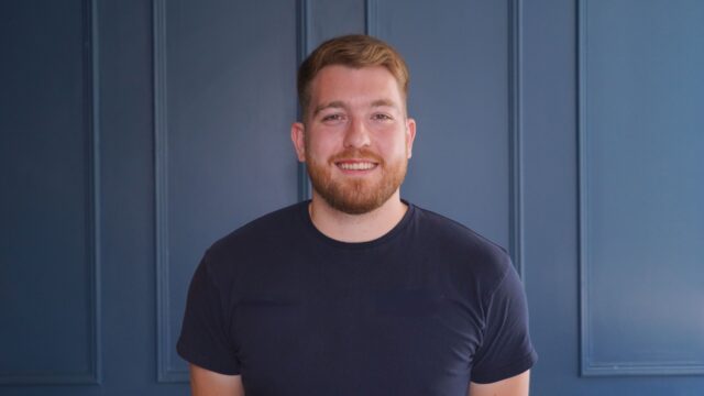 A head and shoulders shot of a ginger man with a beard smiling at the camera