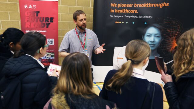 A man in uniform at a careers fair talking to a group of students in front of pull up banners from his company