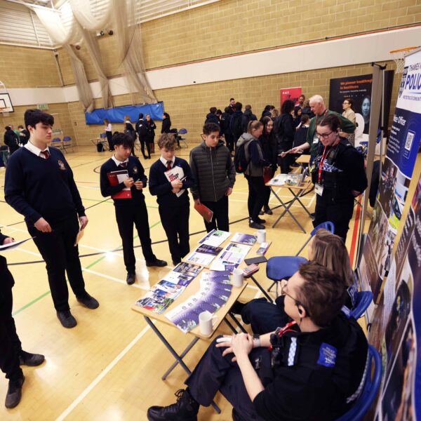 several male students holding booklets that say "get future ready" whilst at a careers fair stall for the police, listening to police offers talk