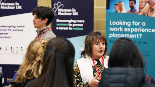 A female employer at a careers fair talking to students. behind her are banners with wording: 'women in nuclear uk' and 'looking for a work experience placement?'