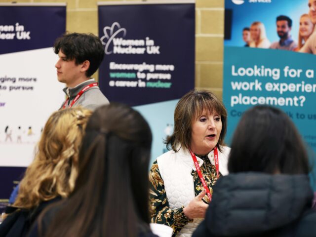 A female employer at a careers fair talking to students. behind her are banners with wording: 'women in nuclear uk' and 'looking for a work experience placement?'