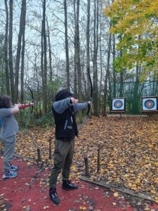 Two people taking part in archery, with these two young people taking shots at specially erected targets on a tree-lined outdoor archery range.