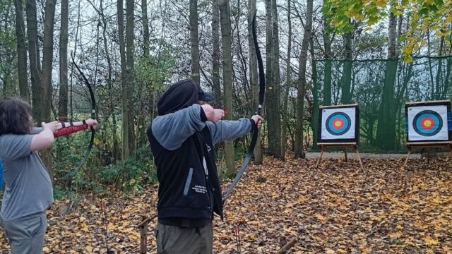 Two people taking part in archery, with these two young people taking shots at specially erected targets on a tree-lined outdoor archery range.