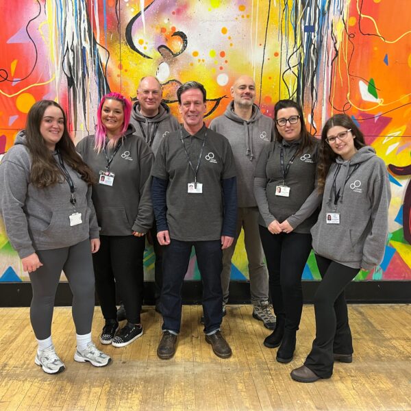 Members of our Liverpool team in a youth centre wearing casual career Connect hoodies and smiling at the camera. Behind them is an orange, yellow and red mural with patterns on,
