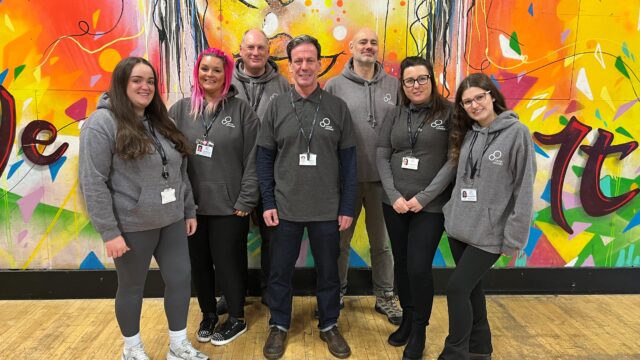 Members of our Liverpool team in a youth centre wearing casual career Connect hoodies and smiling at the camera. Behind them is an orange, yellow and red mural with patterns on,