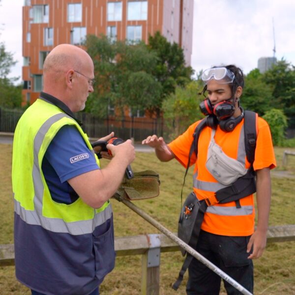 A male young person in high vis and eye protectors, working in a landscaping role. He is smiling and his supervisor is handing him some ear protectors.
