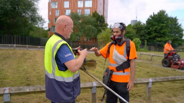 A male young person in high vis and eye protectors, working in a landscaping role. He is smiling and his supervisor is handing him some ear protectors.