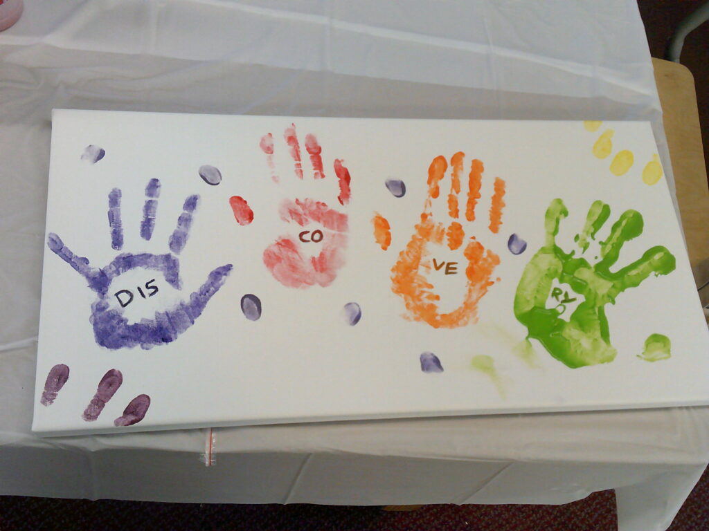 A painting with 4 handprints in different colours and within each hand is letters that spell out "Discovery"