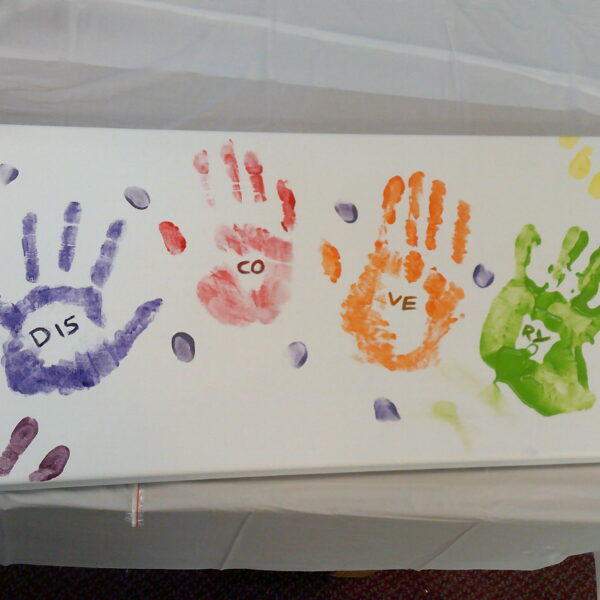 A painting with 4 handprints in different colours and within each hand is letters that spell out "Discovery"