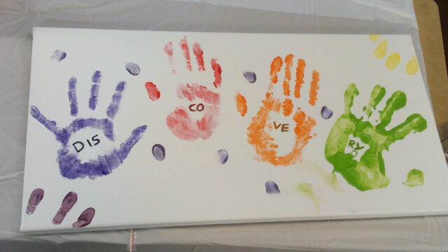 A painting with 4 handprints in different colours and within each hand is letters that spell out "Discovery"