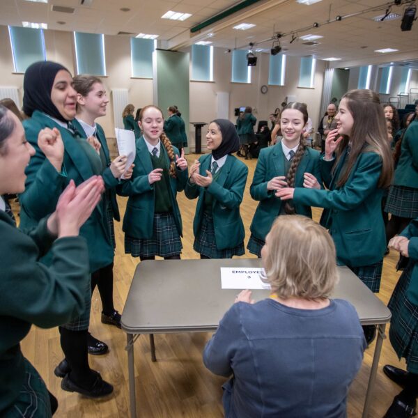 A group of female school students stand around a female employer at a desk, asking them questions to discover what their job is. the pupils are laughing and smiling at the employer.