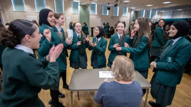 A group of female school students stand around a female employer at a desk, asking them questions to discover what their job is. the pupils are laughing and smiling at the employer.