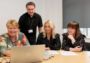 Members of Career Connect's training team delivering a course. Three women are at a desk, with a laptop in front of them. They are explaining course content to a male colleague who is standing up.