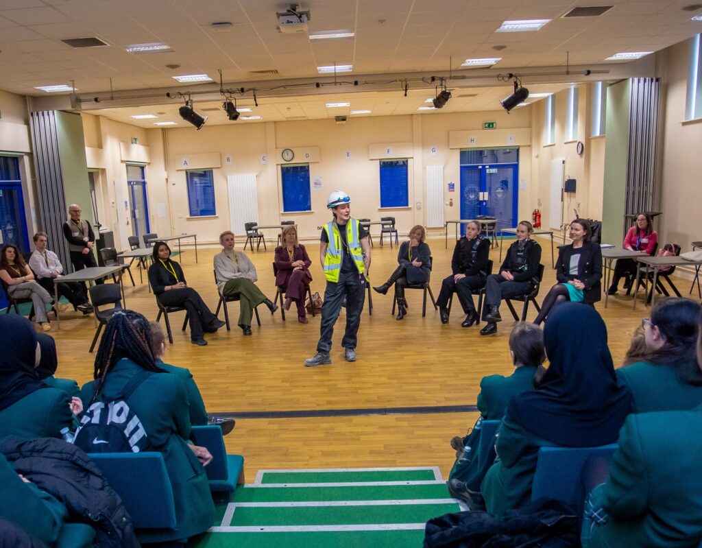 A group of professional women all in different uniforms sat in a horseshoe shape with one woman, who is wearing a hard hat and a hi-vis jacket stood in the middle talking to an audience
