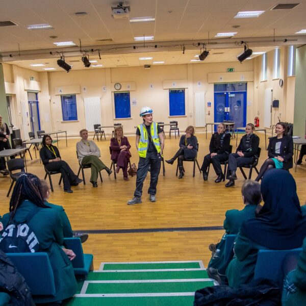 A group of professional women all in different uniforms sat in a horseshoe shape with one woman, who is wearing a hard hat and a hi-vis jacket stood in the middle talking to an audience