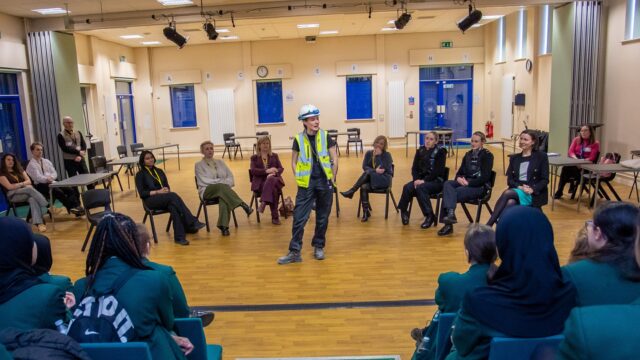 A group of professional women all in different uniforms sat in a horseshoe shape with one woman, who is wearing a hard hat and a hi-vis jacket stood in the middle talking to an audience
