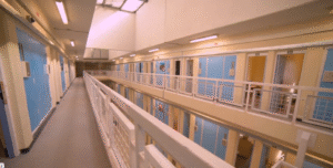The inside of a prison. A shot from the main stairs. Blue cell doors are visible, and the rest of the fittings are cream. It is bright.