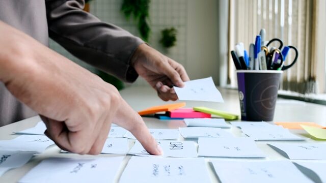 A hand pointing at some sticky notes on a desk with another person's hand in the background holding up a sticky note.