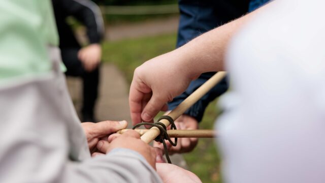 the hands of two people using twine to tie two pieces of wood together.