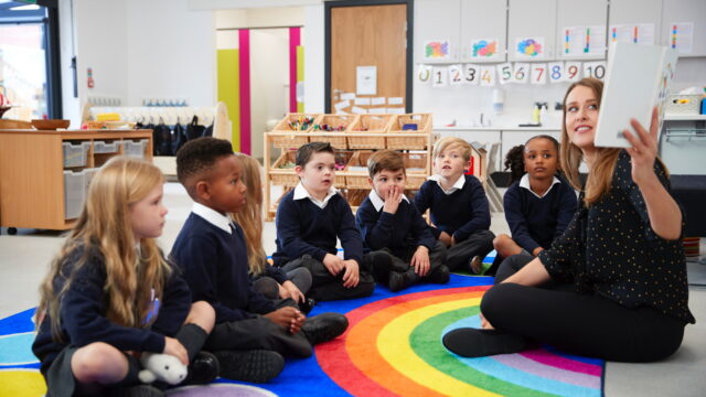 Female teacher holding up a book in front of her class of primary school kids sitting on the floor in a classroom