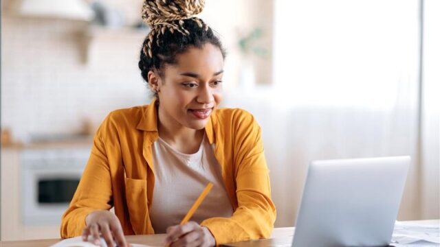 A young woman with dreadlocks up in a bun sitting at a laptop doing an online course. She is looking at the screen and taking notes in a notebook.