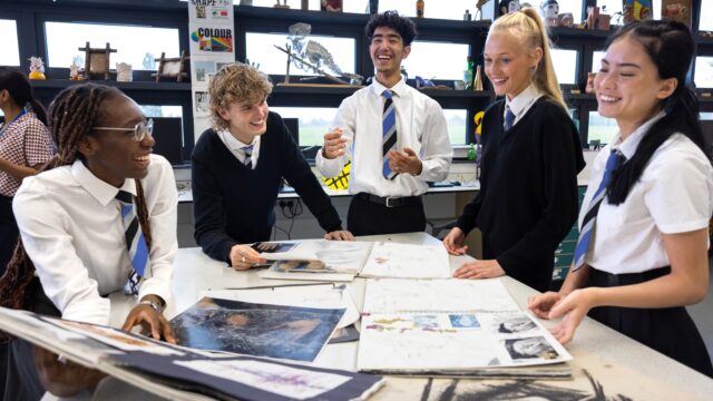 A group of 5 secondary school students stood around a table filled with their work. They are all smiling