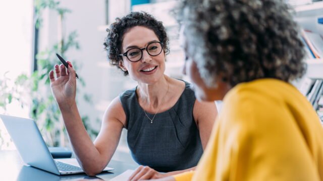 A professional woman sat at a desk holding a pen in her hand and smiling at another woman