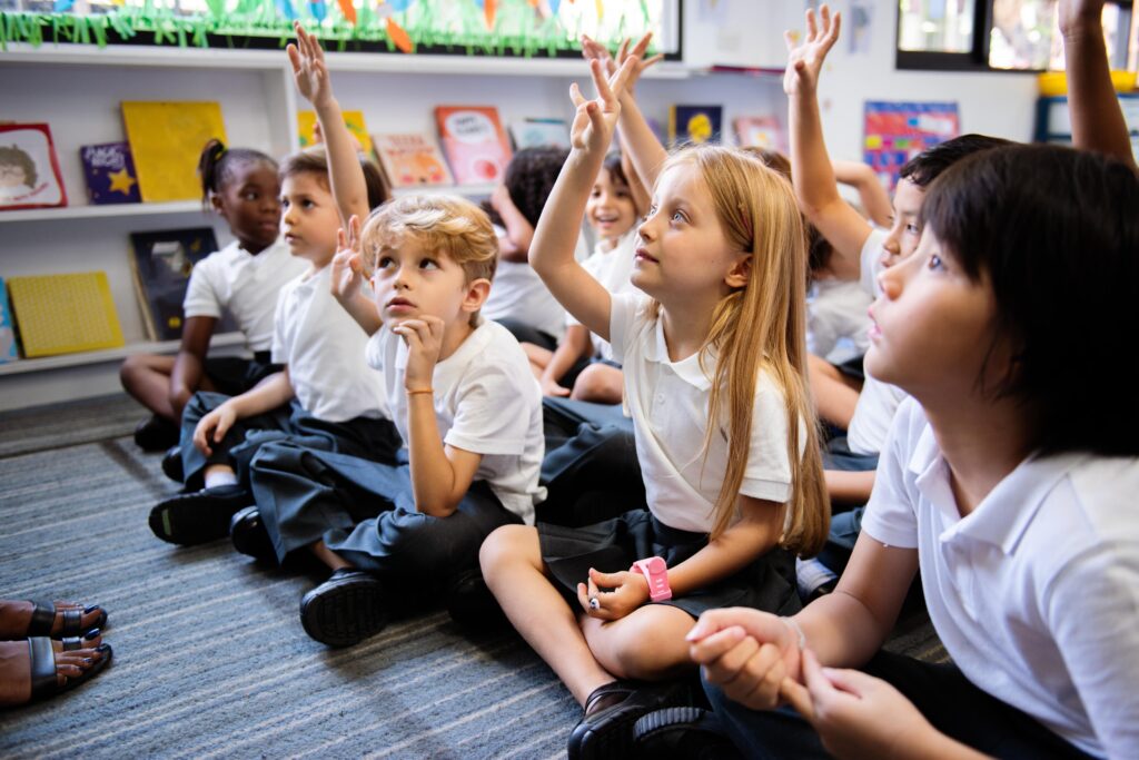 A class of primary school children sat on the floor of a classroom with their hands up to ask a question