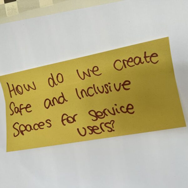 A yellow post-it note on a wall with handwritten text: "How do we create safe an inclusive spaces for service users?"