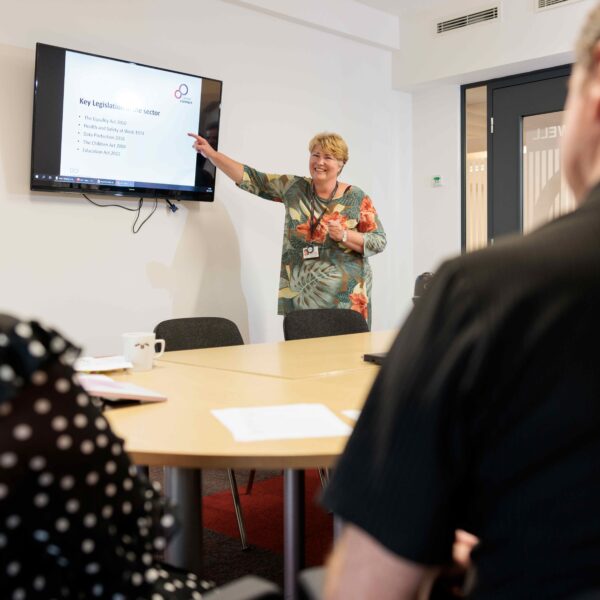 A female member of Career Connect's training team delivering a session. She is stood at the front of the room pointing to a screen with the words 'key legislation'. She is looking into the room and smiling.