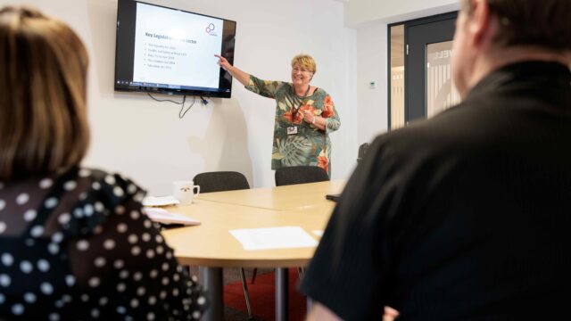 A female member of Career Connect's training team delivering a session. She is stood at the front of the room pointing to a screen with the words 'key legislation'. She is looking into the room and smiling.