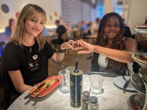 Two female youth ambassadors, making a heart shape with their hands together. They are eating lunch as part of an enrichment activity.