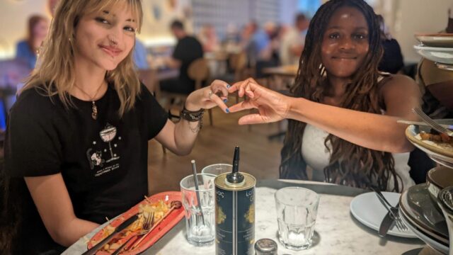 Two female youth ambassadors, making a heart shape with their hands together. They are eating lunch as part of an enrichment activity.