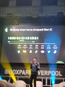 Joe Hall from channel 4 on stage at Raise Up. Behind him is a large screen with a timeline that says 'Events that have shaped Gen Z'