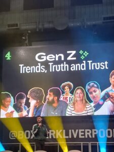 A male speaker - Joe Hall, Senior Research Executive at Channel 4 - is on stage at the Raise Up Event, holding a microphone and speaking. Behind him is a screen with images of young people and the words 'Gen Z - Trends, Truth and Trust'