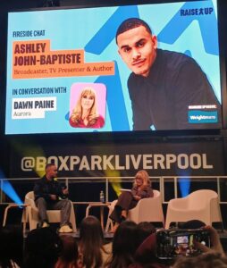 Ashley John Baptiste on stage at Raise Up, sat down in conversation with a female presenter. The screen reads 'fireside chat'
